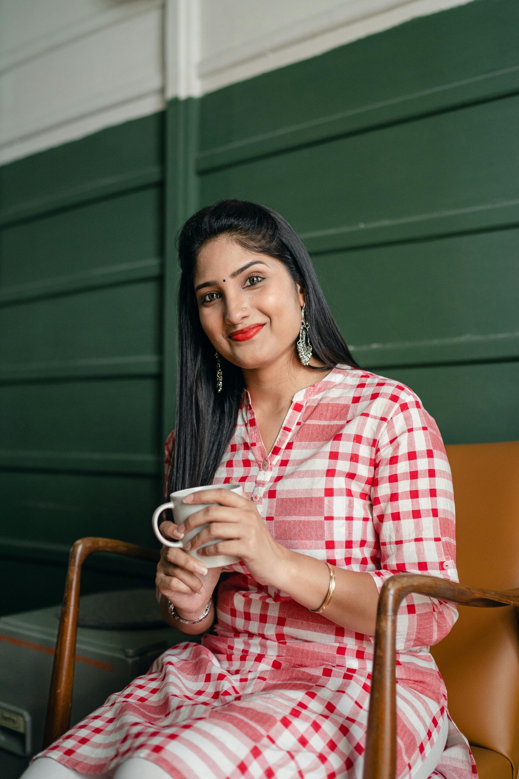 Positive smiling Indian lady in stylish traditional wear sitting on chair against green wall with cup of hot drink and looking at camera